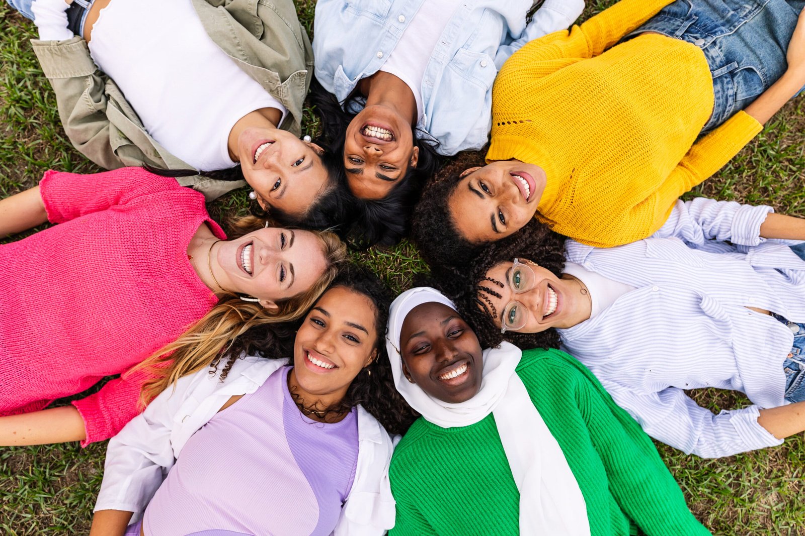 High angle view. Portrait of diverse happy women looking at camera lying together on grass at city park. Happiness, female friendship and youth community concept.