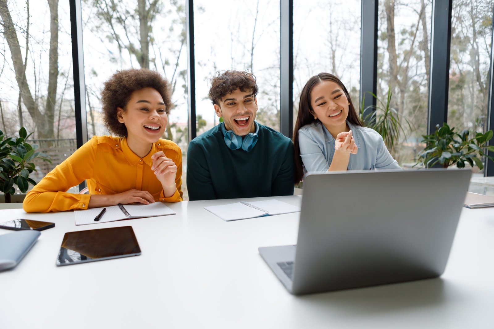 Three cheerful diverse students in casual wear are engrossed and laughing as they look at laptop screen in bright, modern study space with window view